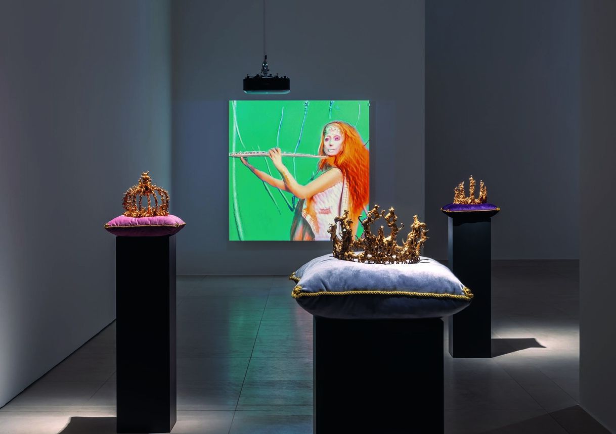 Installation view of Marianna Simnett: Headless, gold crowns on velvet cushions before a projected video